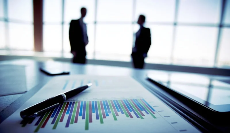 Blurry silhouettes of two people stand by large windows. A table in the foreground has a colorful bar chart and a pen, suggesting a business setting.