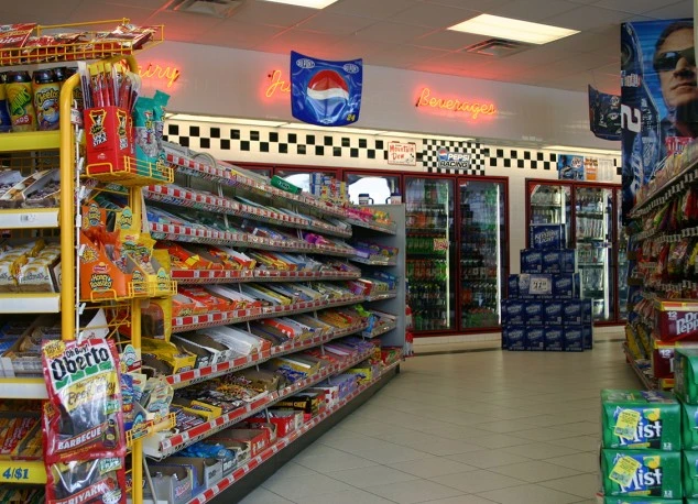 Inside a convenience store, featuring shelves stocked with snacks and candies. Beverages are visible in refrigerators. Racing-themed decor adds to the ambiance.