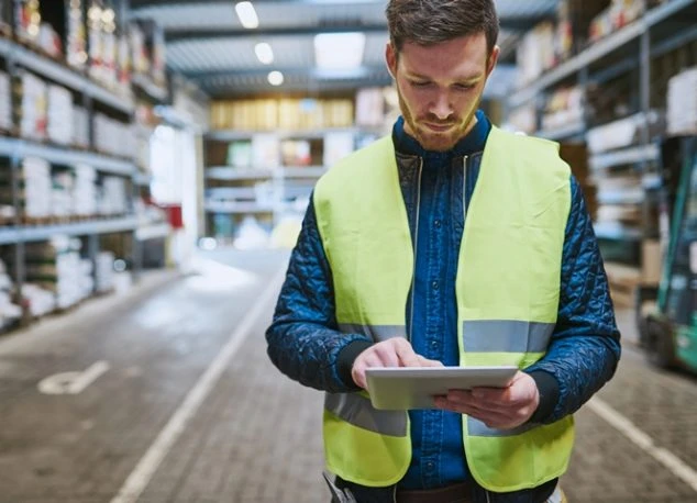 Trade program management in action: warehouse worker wearing a high-visibility safety vest uses a tablet in a warehouse.